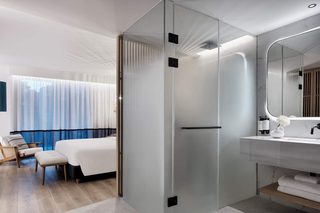 A modern hotel room with a glass partition, a bed, a sitting area with a chair, a large mirror, and a bathroom with a sink and towel rack.