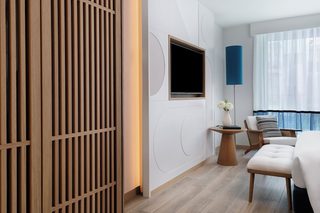 A modern hotel room with a wooden partition, a flat-screen TV mounted on the wall, a round wooden side table with a vase of flowers, a cushioned chair, and a bed with a white duvet.