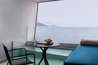A balcony with a glass table, a chair, a bowl of fruit, and a view of the sea.