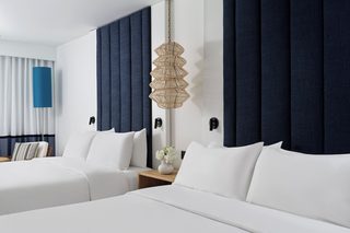 A hotel room with two beds, a large window with curtains, a wall-mounted lamp, and a small table with a vase.
