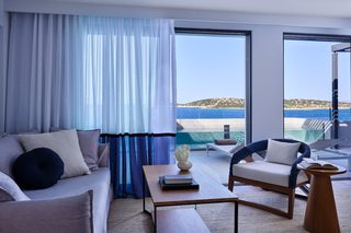 A modern hotel room with a large window offering a view of the sea and a beach. There is a sofa, a chair, and a small table with a book and a vase.