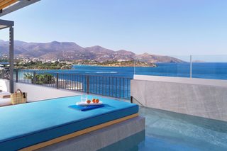 A pool area with a view of the sea and mountains, a lounge chair, and a table with a drink and snacks.