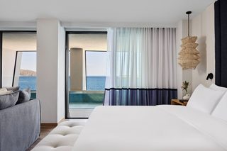 A modern hotel room with a large bed, a sofa, a view of the sea, and a wicker light fixture.