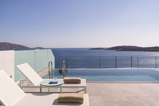 A poolside area with lounge chairs and a table, overlooking a clear blue sea and distant hills.