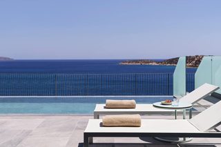 A poolside area with lounge chairs and towels, overlooking a clear blue sea and distant land.