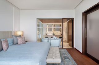 A modern bedroom with a large bed, a bench, a bedside table, a lamp, a bathroom with a bathtub, a mirror, and a door.