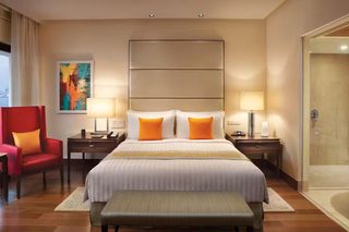 A hotel room with a large bed, two bedside tables, a red armchair, a painting on the wall, and a bathroom door.