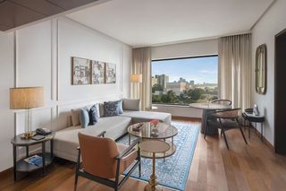 A modern hotel room with a sofa, armchair, round coffee table, and a view of the cityscape through large windows.