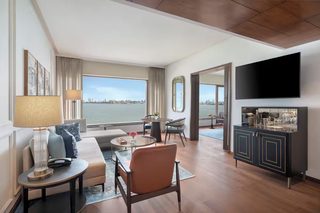 A modern hotel room with a large window offering a city view, a flat-screen TV mounted on the wall, a dining area with a table and chairs, and a bar area with a mirror and glassware.