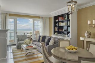 A modern living room with a sofa, armchairs, a coffee table, and a dining table. There is a bookshelf with books and decorative items. A large window offers a view of the ocean.