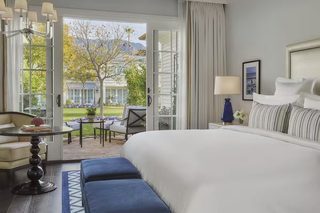 A hotel room with a large bed, a dining area with a table and chairs, and a view of a building and trees outside.