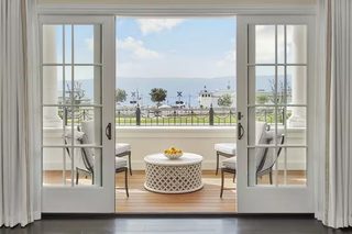 A room with a view through a glass door, featuring a round table with a bowl of fruit, two chairs, and a view of a park and distant mountains.