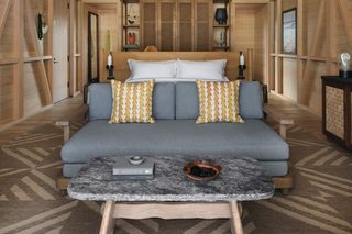 A cozy room with a wooden ceiling, a grey sofa with patterned cushions, a small table with a bowl and books, and a wooden cabinet.
