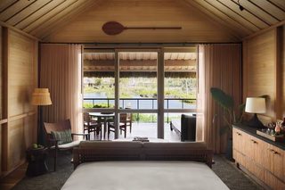 A room with a large bed, a dining table with chairs, and a view of a water body and greenery outside.
