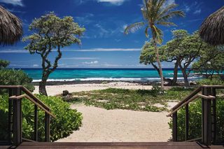A beach view with a wooden staircase leading to a sandy path, surrounded by trees and thatched-roof structures.