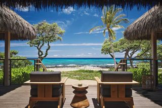A wooden deck with two lounge chairs facing a beach with clear turquoise water and palm trees.